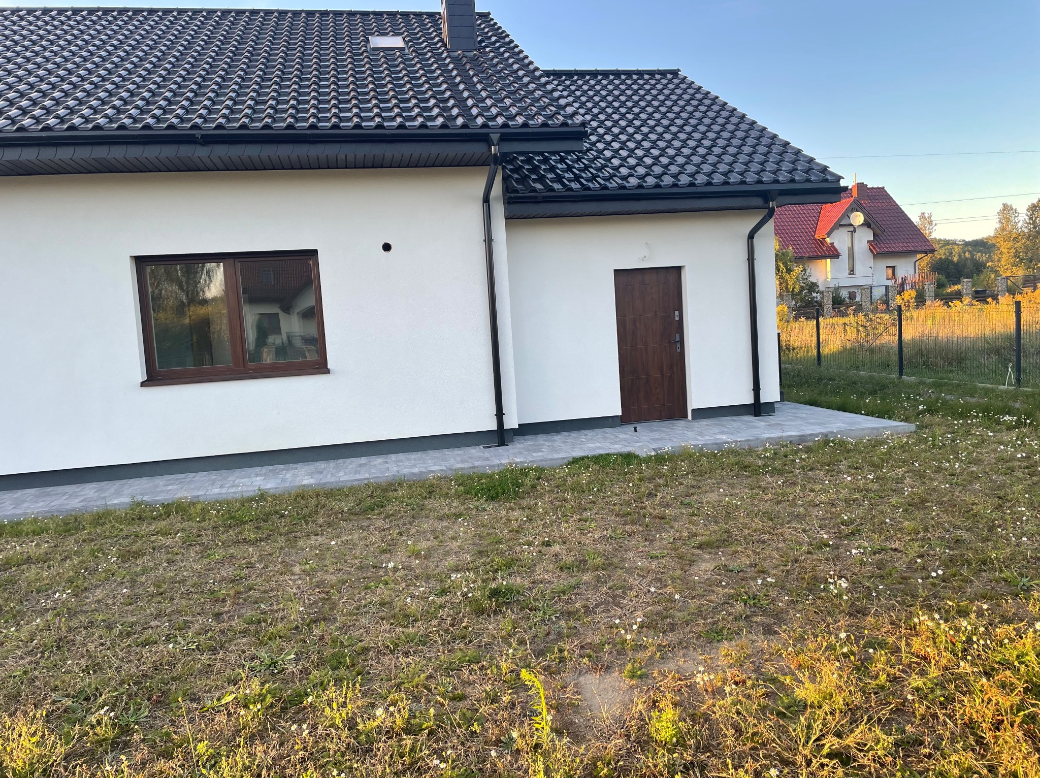 Nowo otynkowany, biały dom jednorodzinny z brązowymi oknami i drzwiami, wykończony ciemną dachówką i szarą kostką brukową wokół budynku, w tle widoczny inny dom z czerwoną dachówką.