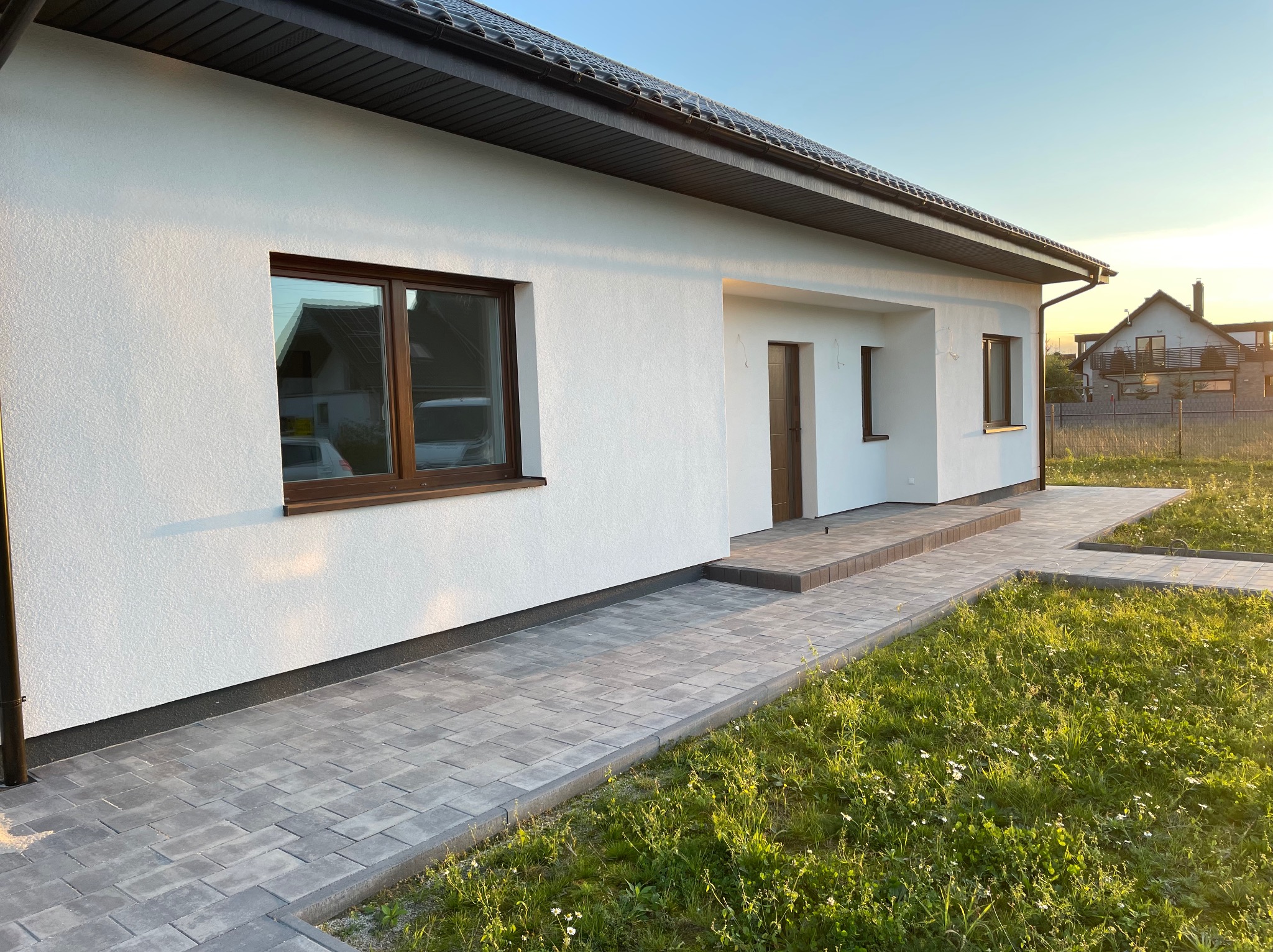 Biała elewacja domu jednorodzinnego z brązowymi oknami i ciemną dachówką, widok z boku na ścieżkę z kostki brukowej w promieniach zachodzącego słońca.