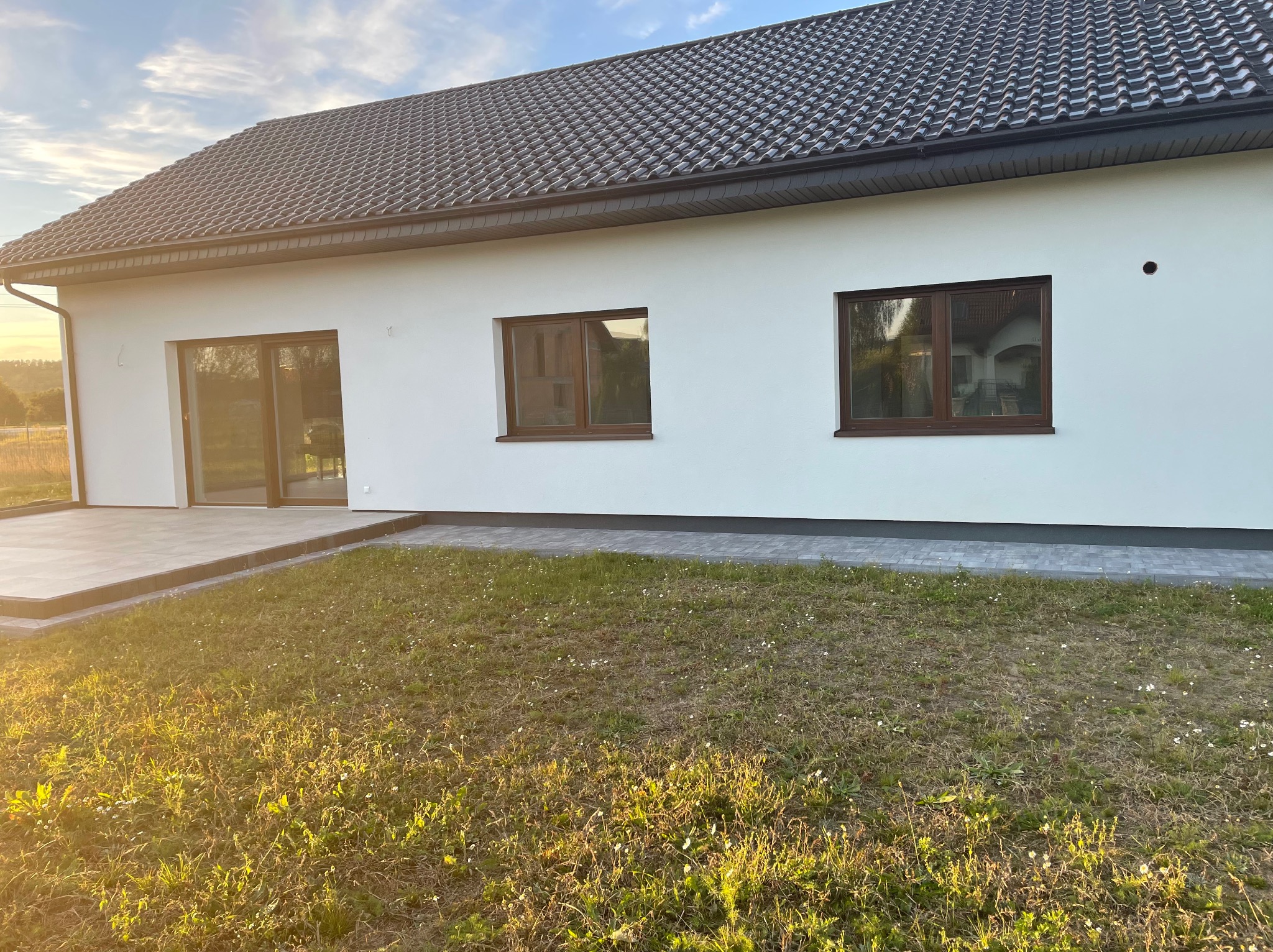 Nowoczesny dom jednorodzinny z białymi ścianami, ciemną dachówką i brązowymi ramami okiennymi, widok z zewnątrz na trawnik w promieniach zachodzącego słońca.