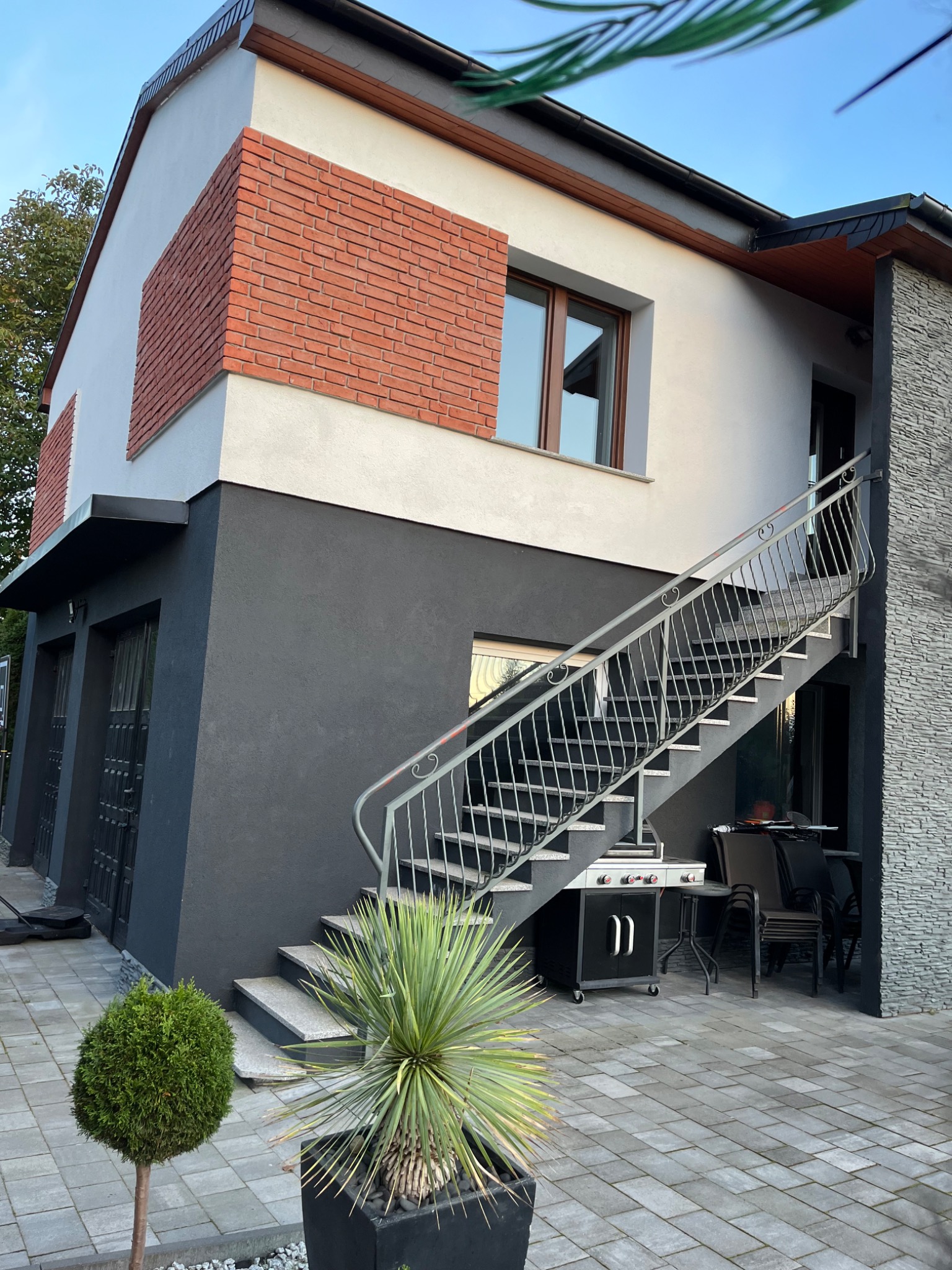 Dwupiętrowy dom z ocieploną elewacją w kolorach bieli i grafitu, z dekoracyjnym fragmentem z czerwonej cegły, schody zewnętrzne z metalową balustradą i kostka brukowa na podwórku.