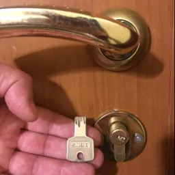 Otw&oacute;r zamka w drewnianych drzwiach i klucz ABUS trzymany w dłoni, złota klamka powyżej.