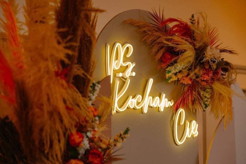 Neonowy napis 'My się kochamy' otoczony kompozycją z suszonych traw i kwiatów w ciepłych barwach, oświetlony miękkim światłem.