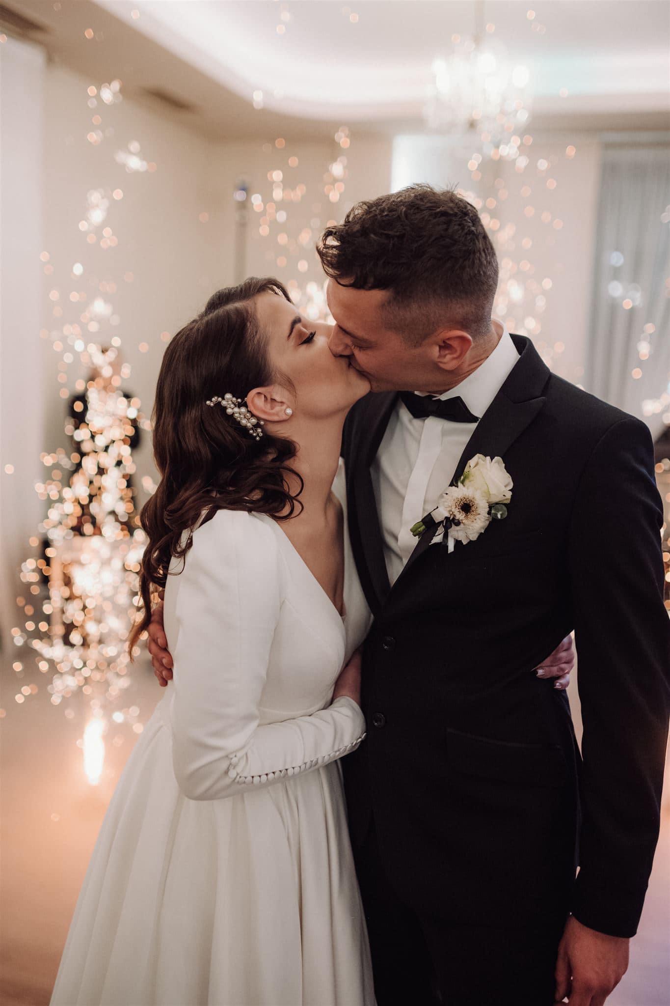 Pocałunek nowożeńców na tle zimnych ogni podczas wesela; panna młoda z ozdobą z pereł we włosach, pan młody w czarnym smokingu z butonierką.