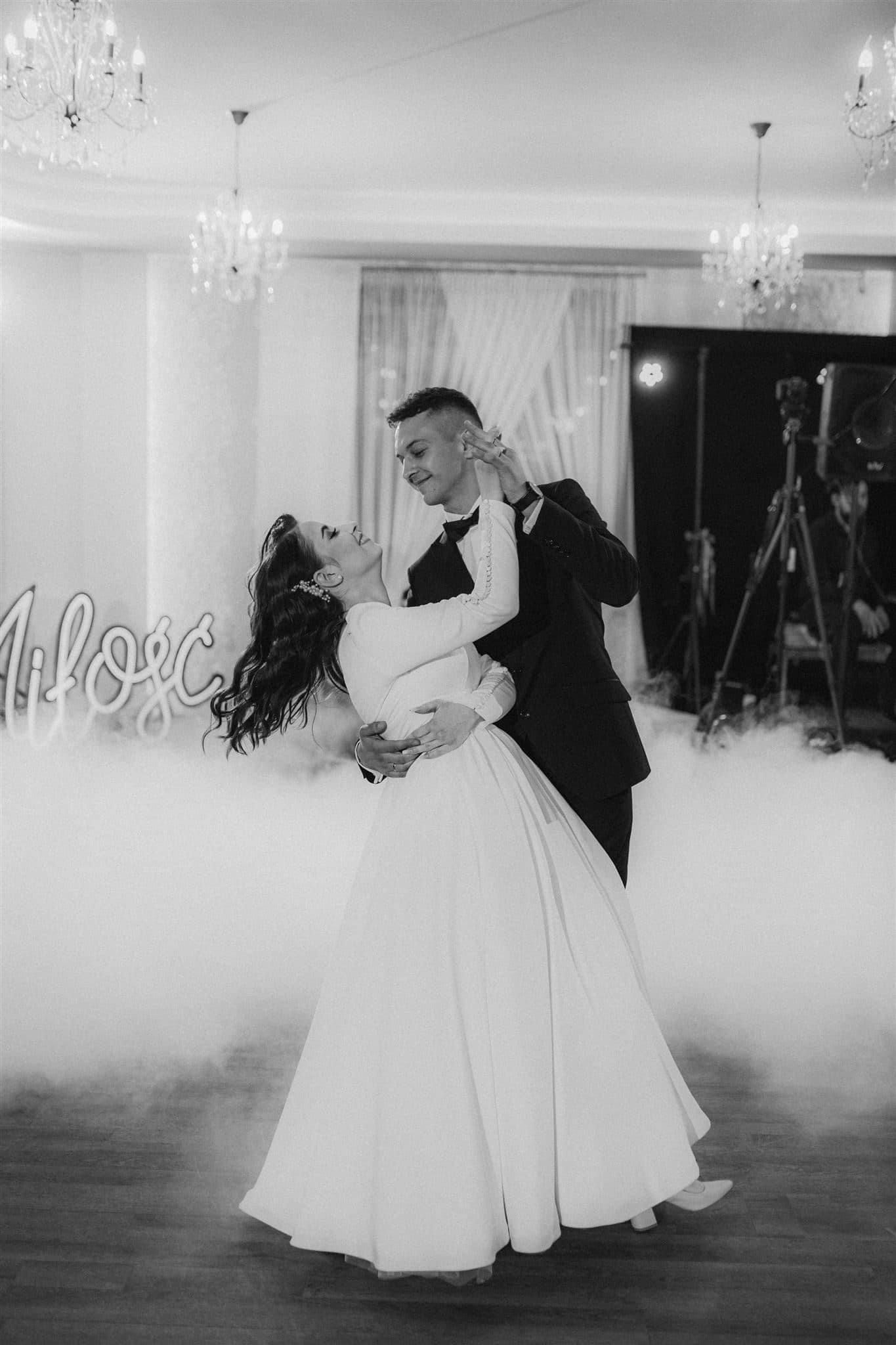 Czarno-białe zdjęcie pary młodej tańczącej w dymie, widoczny neon 'Miłość' w tle, dekoracja sali z żyrandolami.