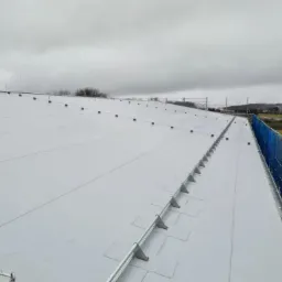 Biały dach przemysłowy z zabezpieczeniami przeciwśniegowymi i siatką ochronną, widok z góry, pochmurne niebo.