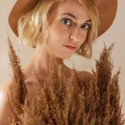 Portret kobiety w brązowym kapeluszu, częściowo zasłoniętej przez pampasową trawę, z delikatnym uśmiechem i spojrzeniem w obiektyw.