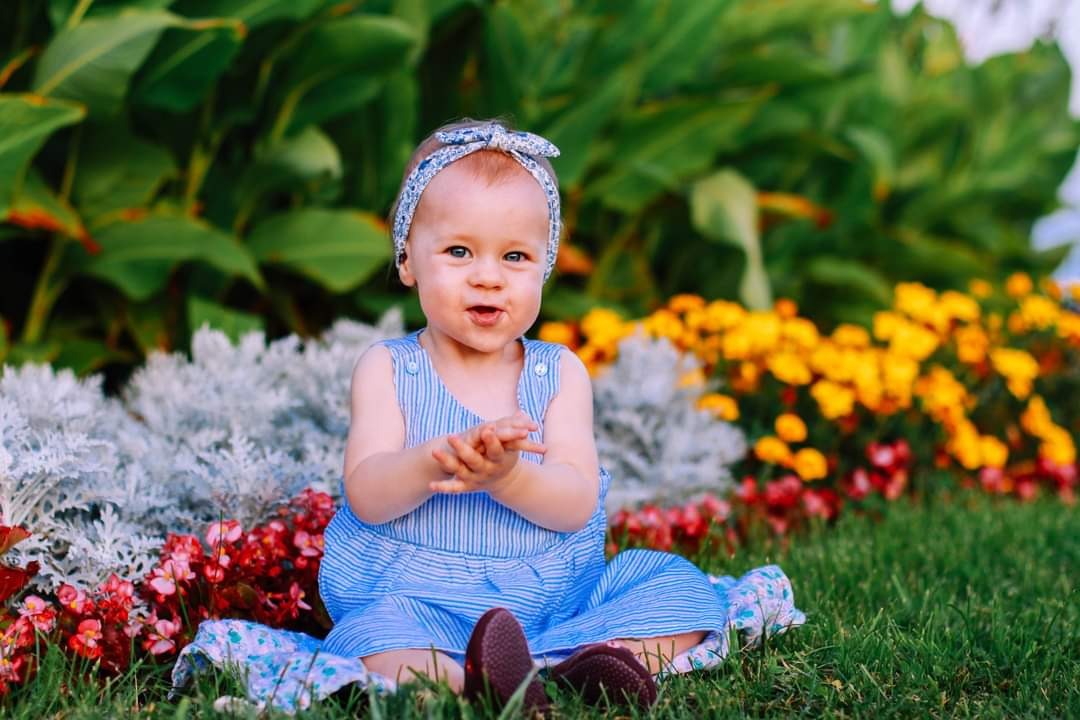 Urocza roczna dziewczynka w niebieskiej sukience w paski i opasce siedzi na trawie przed kolorowymi kwiatami, z dłońmi złożonymi jakby klaskała.