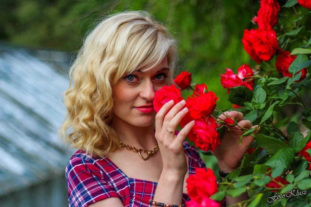 Portret kobiety z blond kręconymi włosami w czerwonej kraciastej sukience, pozującej z bukietem czerwonych róż na tle zieleni.