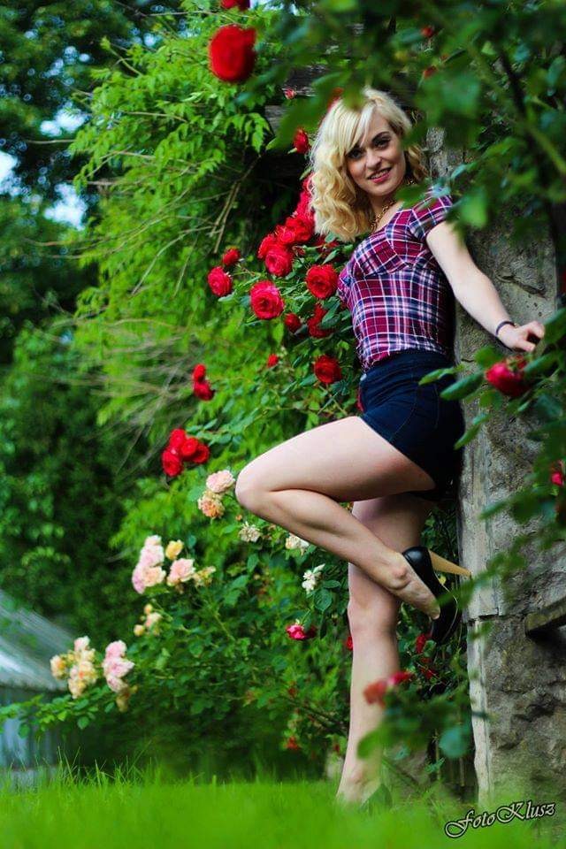 Uśmiechnięta blondynka w kraciastej koszuli i czarnych szortach opiera się o kamienny murek porośnięty różami w ogrodzie, pozując na jednej nodze w czarnych szpilkach.