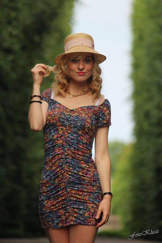 Portret kobiety w kwiecistej sukience i słomkowym kapeluszu na tle zieleni alei parkowej, z delikatnym uśmiechem i dłonią bawiącą się kosmykiem włosów.