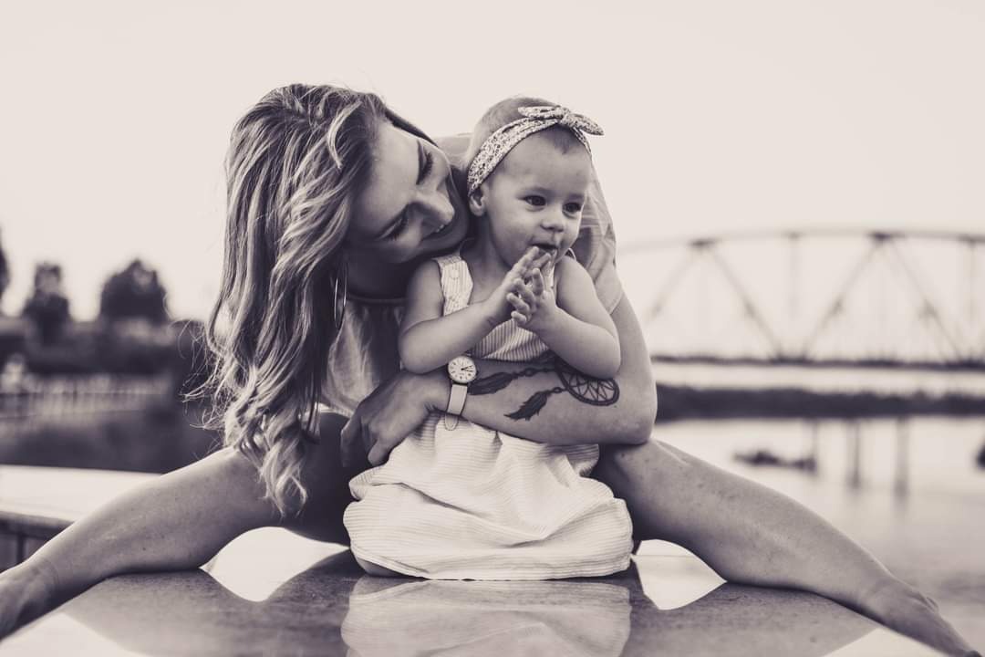 Czarno-biała fotografia matki trzymającej na kolanach małą dziewczynkę w sukience z opaską na głowie, w tle most na rzece w Tczewie, widoczny tatuaż na ramieniu kobiety.