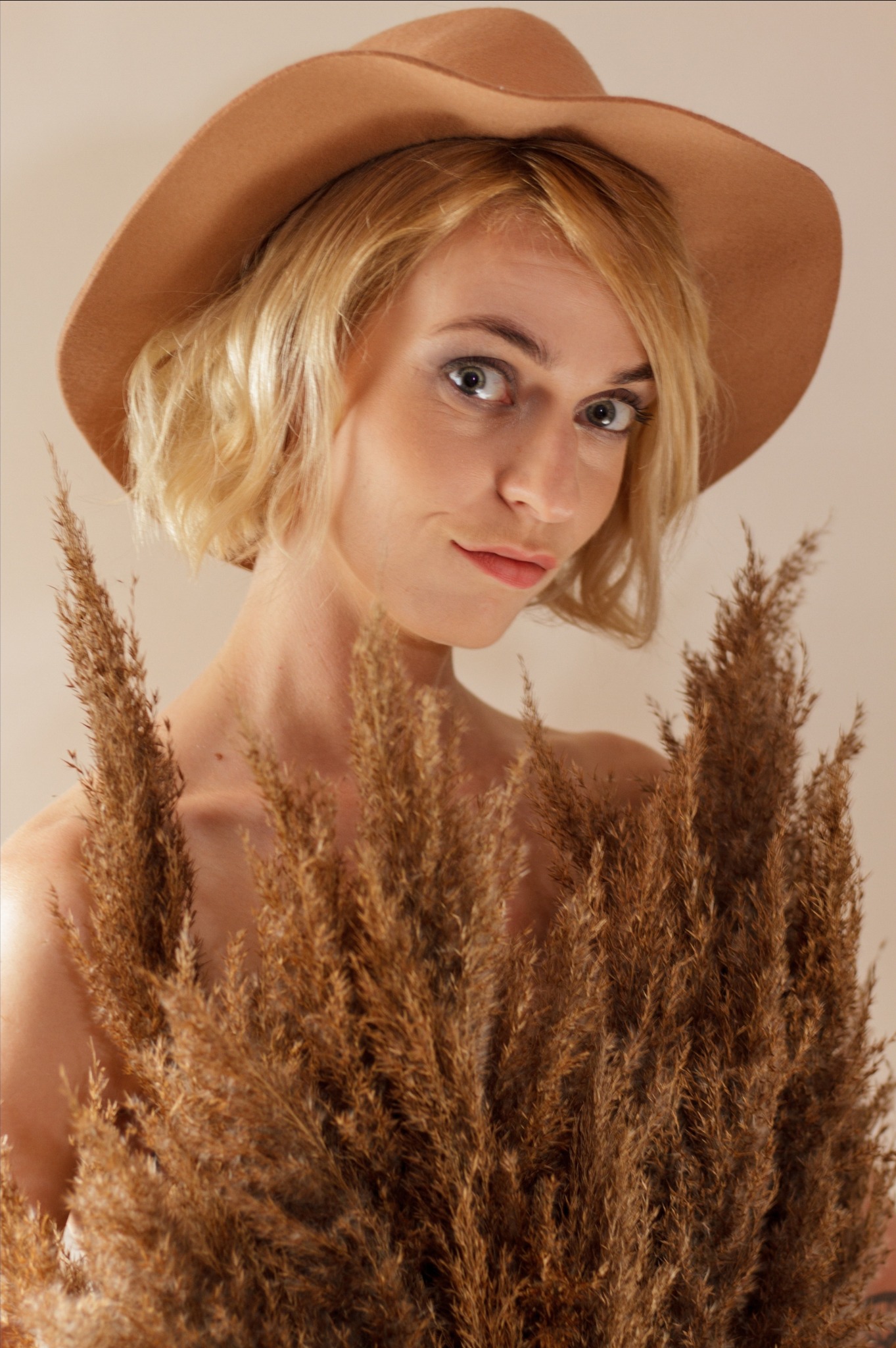 Portret kobiety w brązowym kapeluszu, częściowo zasłoniętej przez pampasową trawę, z delikatnym uśmiechem i spojrzeniem w obiektyw.