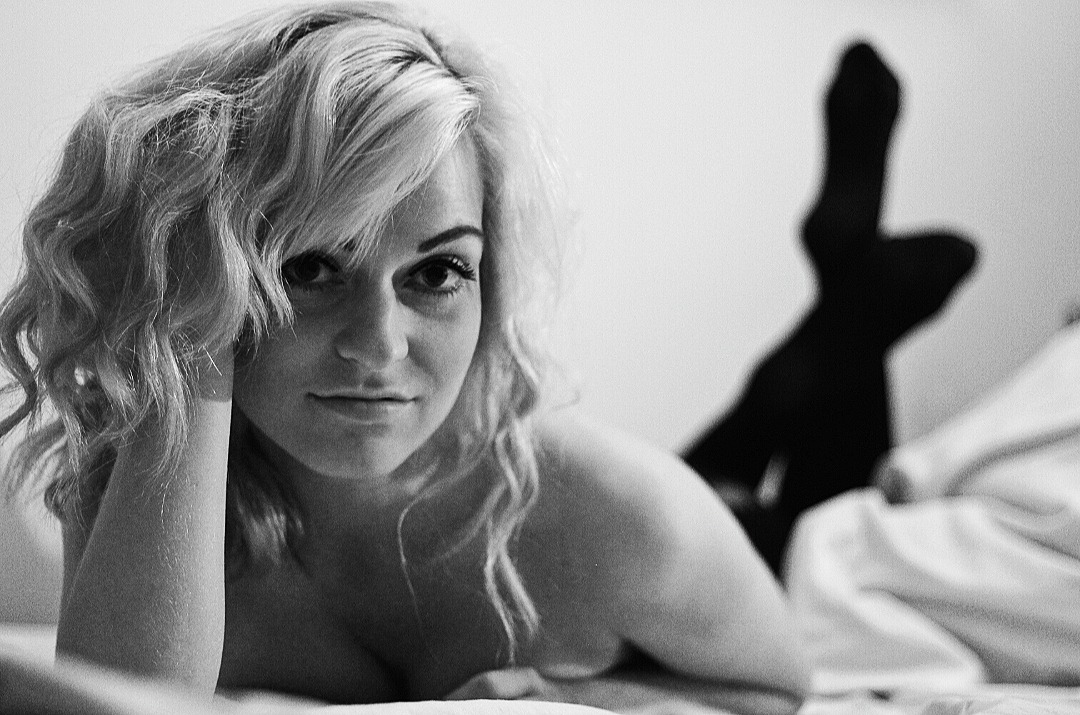 Portret kobiety z blond włosami leżącej na łóżku, w tle widoczne nogi w czarnych rajstopach, czarno-biała fotografia.