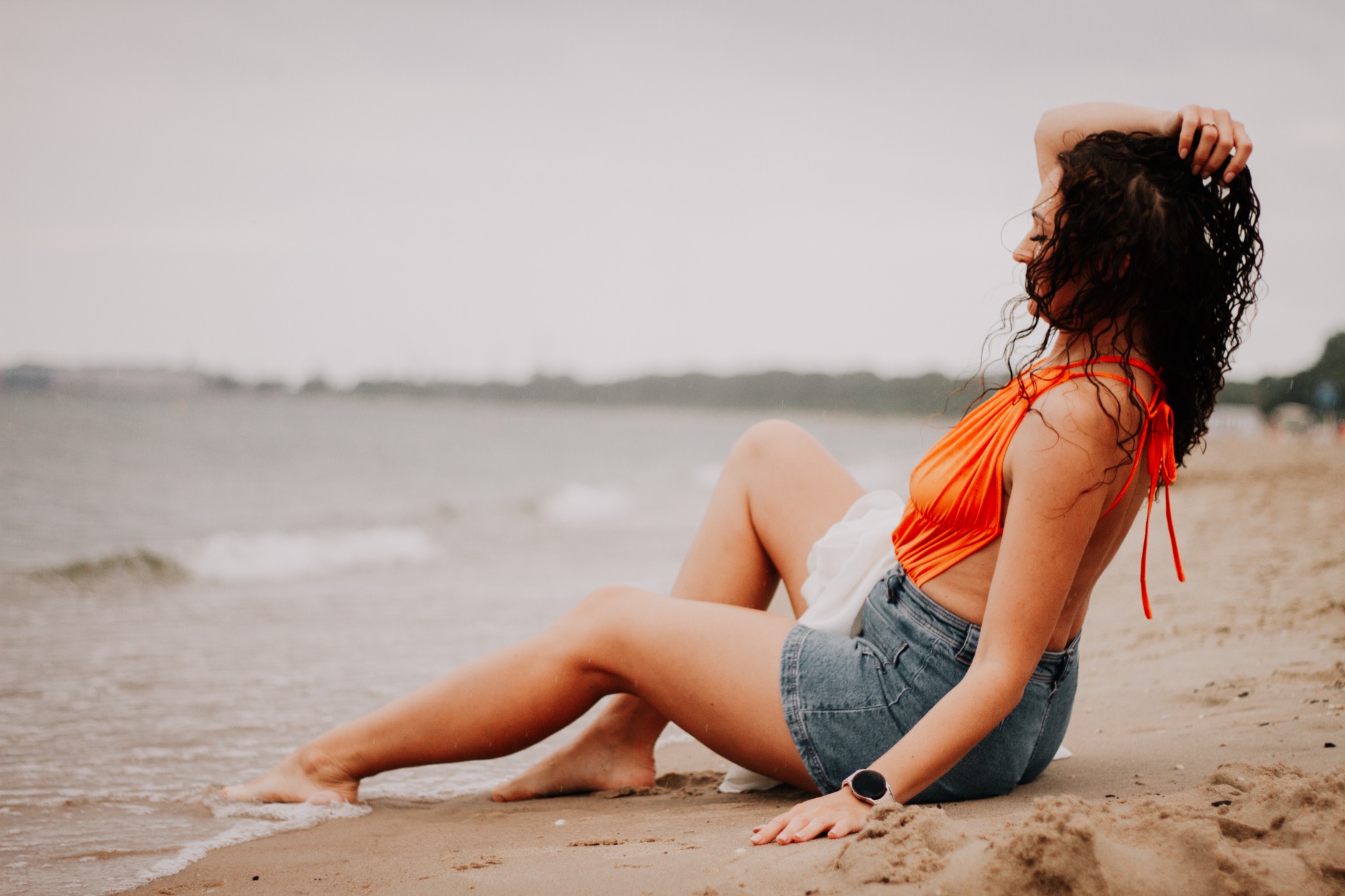 Kobieta w pomarańczowym topie i dżinsowych szortach siedzi na piaszczystej plaży, dotykając kręconych włosów, z widocznym zegarkiem na nadgarstku i falami w tle.