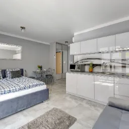 Jasne, nowoczesne studio: łóżko z szarą ramą i niebiesko-białą pościelą, aneks kuchenny z białymi szafkami, szary blat, ananas i napoje, stół z krzesłami, lustro na ścianie, szara podłoga.