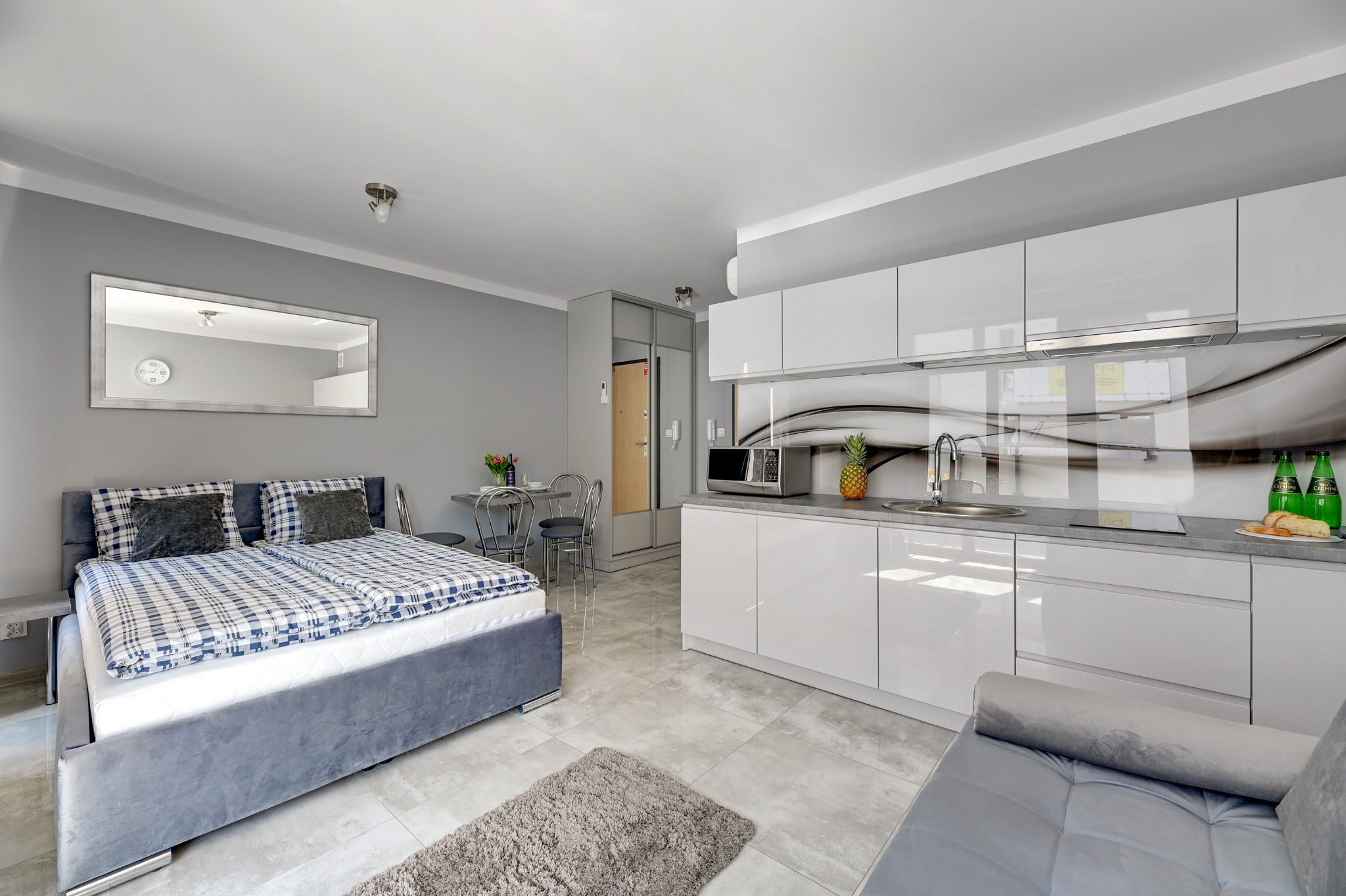 Jasne, nowoczesne studio: łóżko z szarą ramą i niebiesko-białą pościelą, aneks kuchenny z białymi szafkami, szary blat, ananas i napoje, stół z krzesłami, lustro na ścianie, szara podłoga.