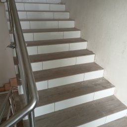 Od kraju do kraju - Schody wykończone płytkami imitującymi drewno z białymi cokołami, widoczna metalowa barierka i karton na górnym biegu schodów.