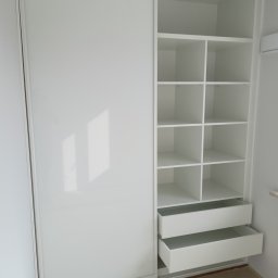 Od kraju do kraju - Biała szafa wnękowa z przesuwnymi drzwiami i otwartymi półkami oraz dwiema wysuniętymi szufladami, widoczne zabezpieczenia podłogi podczas montażu.