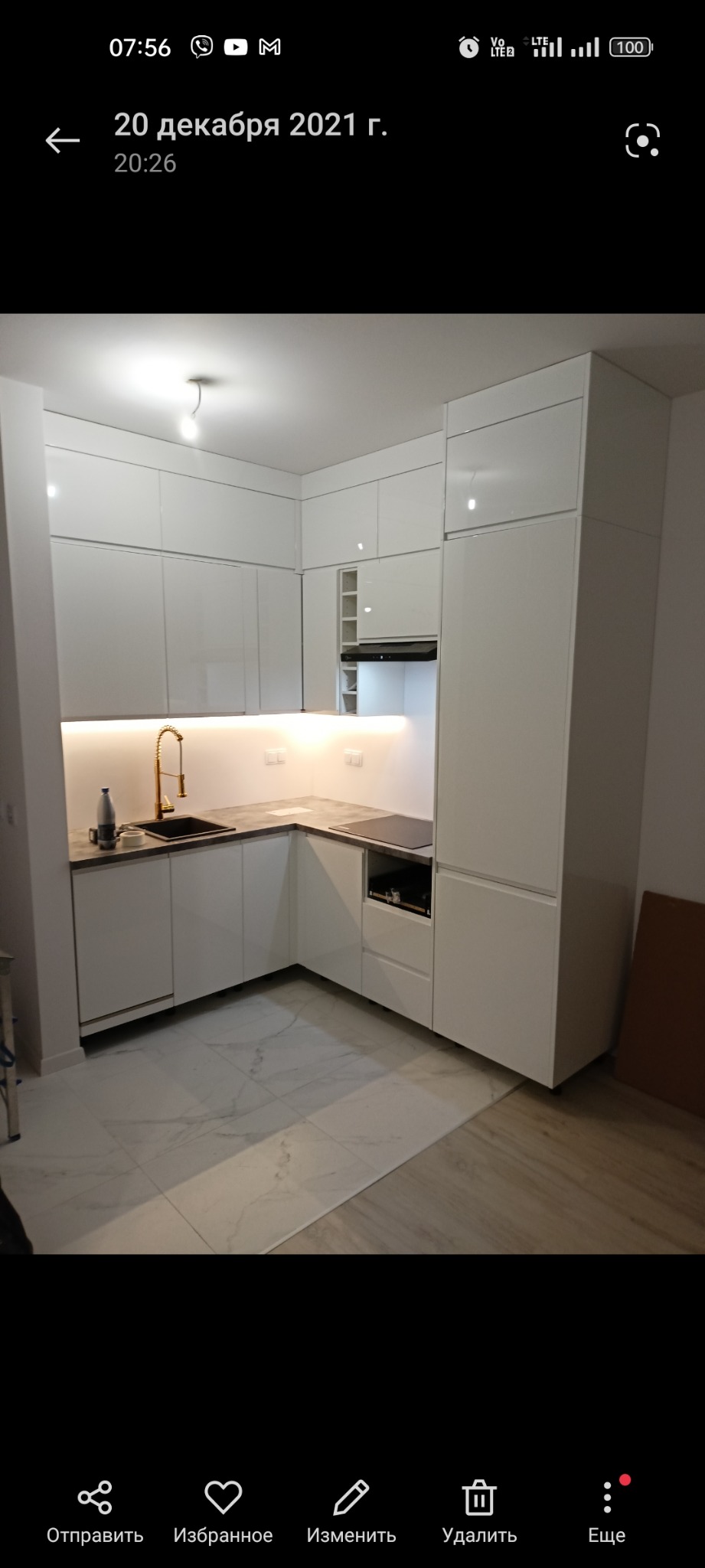 Nowoczesna, biała kuchnia z błyszczącymi frontami, złotą armaturą i podświetleniem LED podszafkowym, marmurowa podłoga, widok na narożnik z blatem w kolorze betonu.