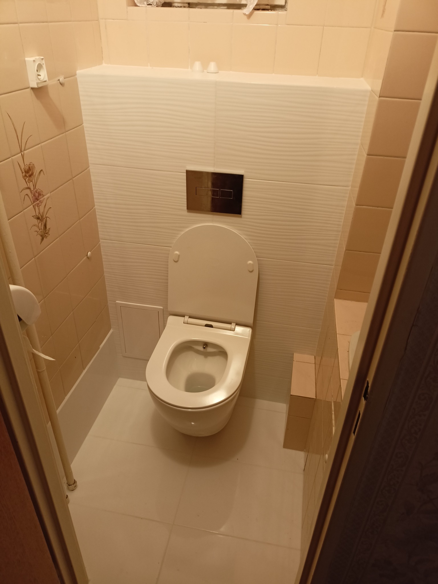 Zamontowana toaleta podwieszana z chromowanym przyciskiem spłukującym na tle beżowych płytek w małej łazience.