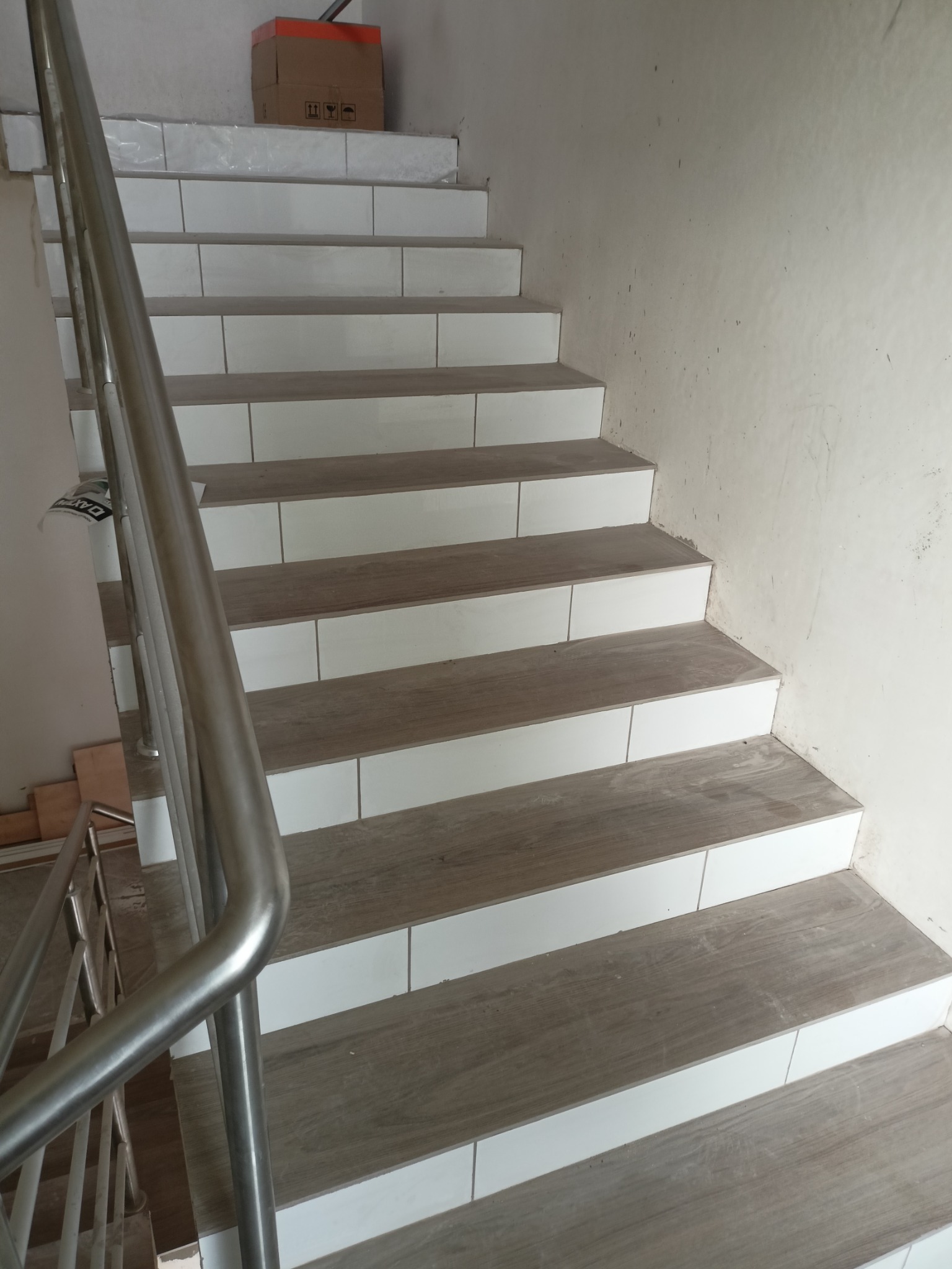 Schody wykończone płytkami imitującymi drewno z białymi cokołami, widoczna metalowa barierka i karton na górnym biegu schodów.