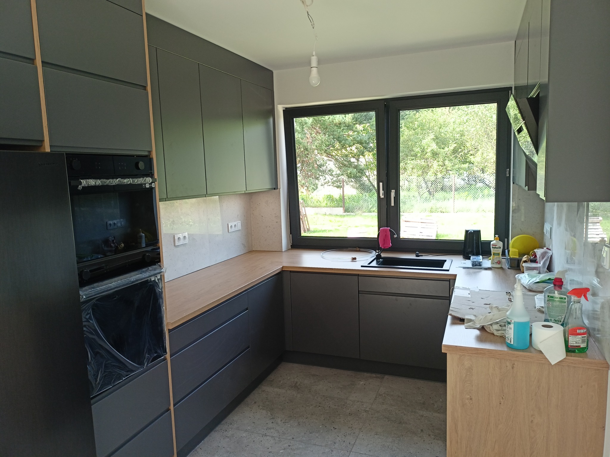 Nowoczesna kuchnia z szarymi szafkami i drewnianym blatem, widok na ogród przez duże okno, sprzęt AGD zabezpieczony folią, środki czystości na blacie.