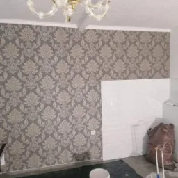 Pokój po położeniu wzorzystej tapety w odcieniach szarości, widoczny fragment ściany wyłożonej białymi, prostokątnymi płytkami, zakryta podłoga, wiadro z farbą i ozdobny żyrandol.