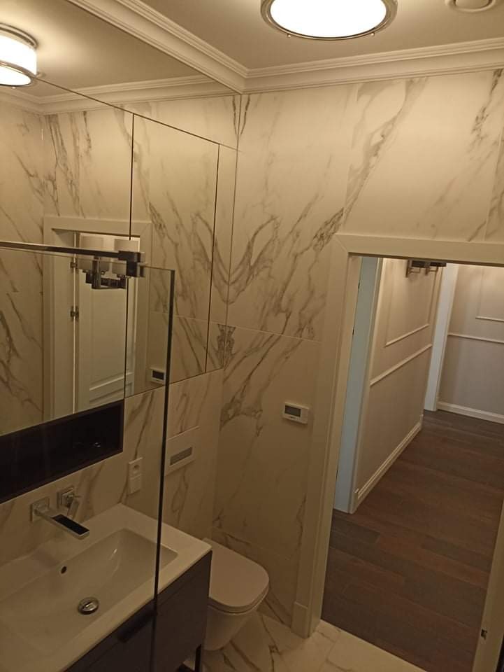 Nowoczesna łazienka z marmurowymi płytkami na ścianach, lustrem nad umywalką i widokiem na korytarz z drewnianą podłogą.