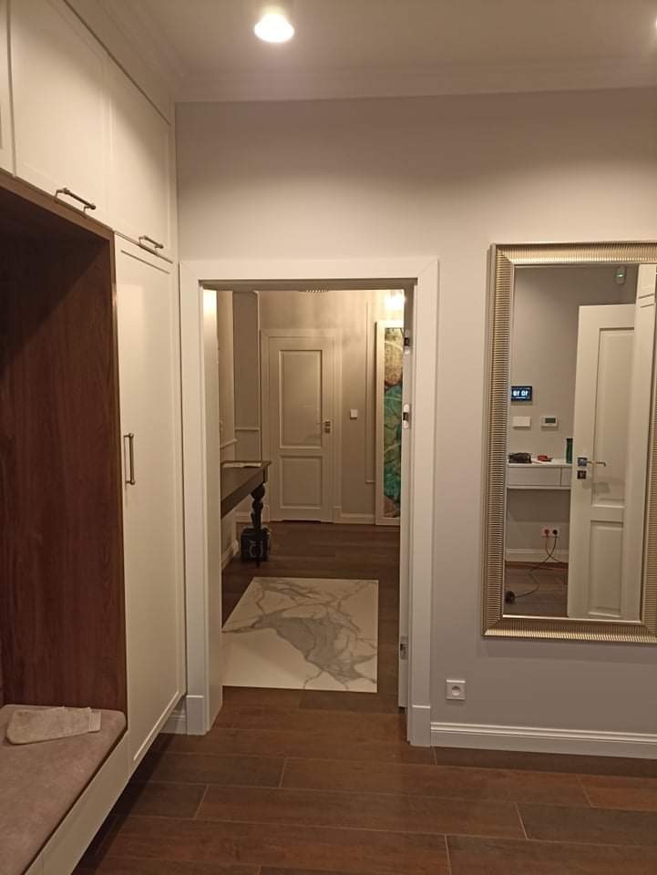 Nowoczesny przedpokój z brązowymi panelami podłogowymi, zabudowaną szafą z siedziskiem, lustrem w złotej ramie i widokiem na dalszą część mieszkania z białymi drzwiami i dywanem z motywem marmuru.