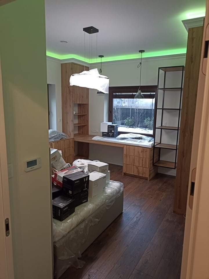 Pokój po remoncie z drewnianą podłogą, biurkiem w zabudowie i regałem, częściowo zagracony kartonami, oświetlony lampami wiszącymi i ledowym paskiem na suficie.