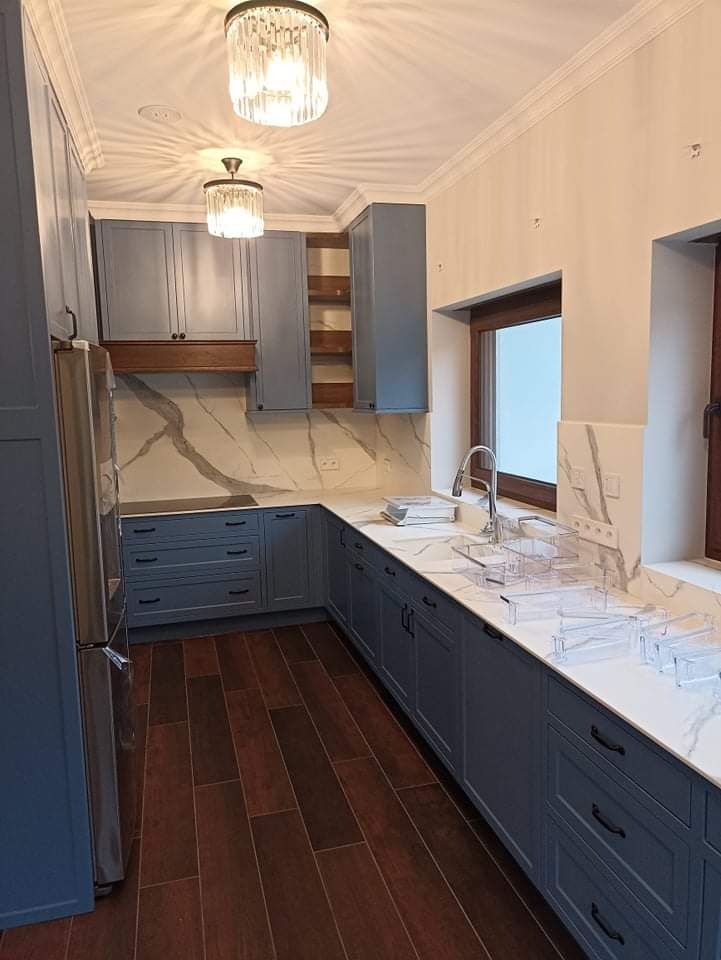 Nowoczesna kuchnia z szarymi szafkami, marmurowym blatem i brązową podłogą; widoczne kryształowe lampy sufitowe i okno z drewnianą ramą.