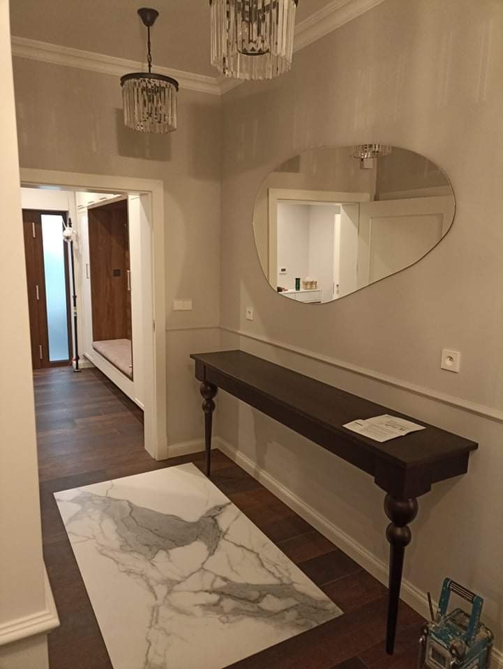 Wnętrze przedpokoju z ciemnobrązową podłogą, biało-szarym dywanem imitującym marmur, długim, wąskim, ciemnym drewnianym stolikiem na toczonych nogach, asymetrycznym lustrem na ścianie i dwoma...