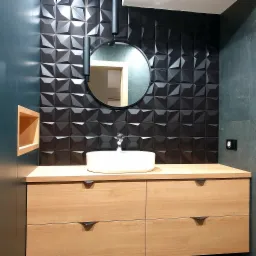 Nowoczesna łazienka z umywalką nablatową, drewnianą szafką z czterema szufladami i czarnymi, trójwymiarowymi płytkami na ścianie, uzupełniona okrągłym lustrem i minimalistyczną lampą.