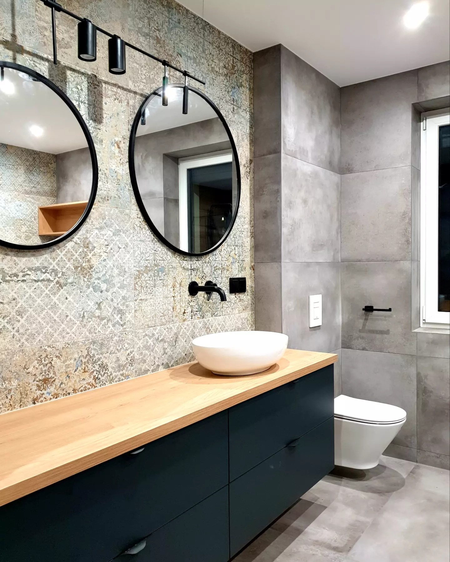 Nowoczesna łazienka z umywalką nablatową, dwiema okrągłymi lustrami z czarną ramą i wzorzystymi płytkami na ścianie, oświetlona reflektorami szynowymi.
