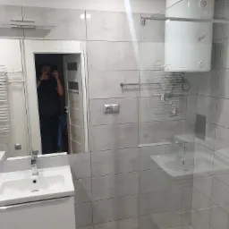 Wyremontowana łazienka z szarymi płytkami imitującymi beton, kabiną prysznicową ze szklaną ścianką, białym bojlerem oraz odbiciem fotografa w lustrze nad umywalką.