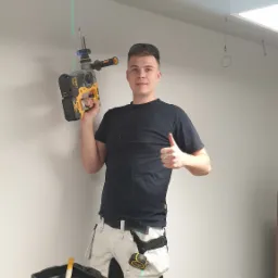 Uśmiechnięty monter na drabinie z wiertarką DeWalt z laserem krzyżowym celuje w sufit z klimatyzacją kasetonową, ubrany w robocze spodnie Snickers z narzędziami w kieszeniach i czarną koszulkę.