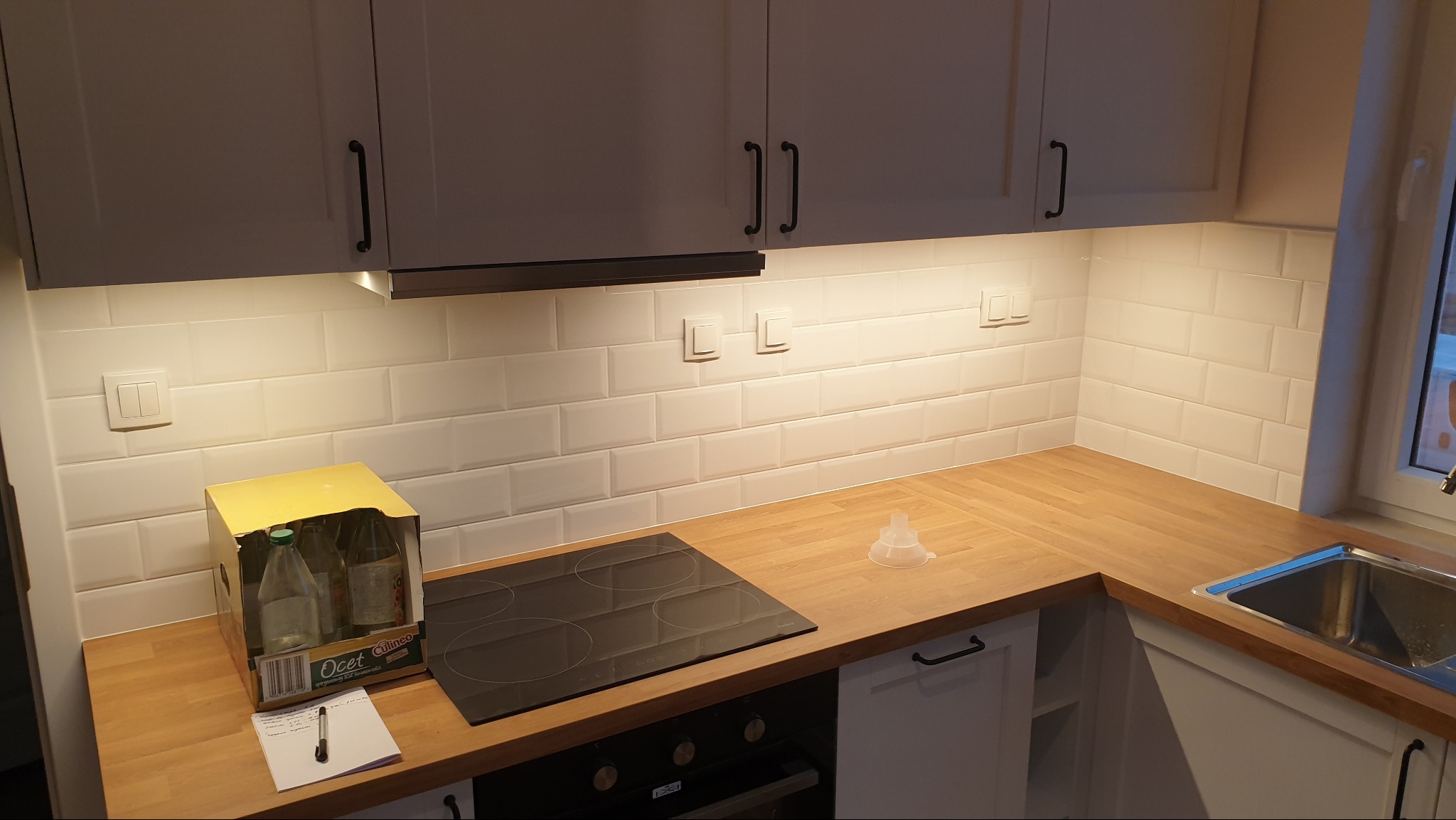 Nowoczesna kuchnia z białymi szafkami, drewnianym blatem i białymi płytkami na ścianie. Widoczna płyta indukcyjna, zlewozmywak i pudełko z octem na blacie.