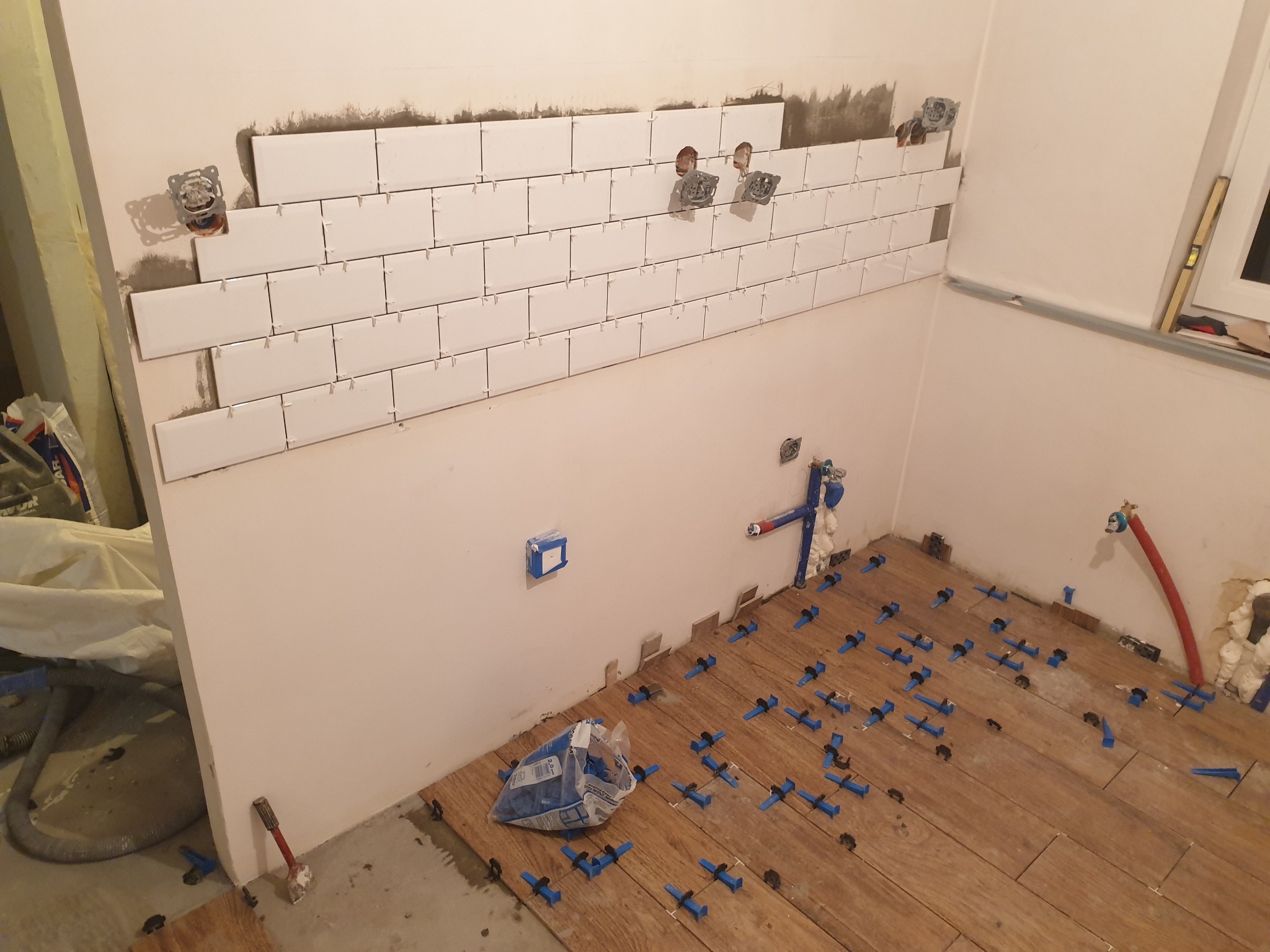 Białe, prostokątne płytki częściowo ułożone na ścianie z przygotowanymi otworami na gniazdka elektryczne; na podłodze drewnopodobne panele z niebieskimi krzyżykami dystansowymi i workiem...