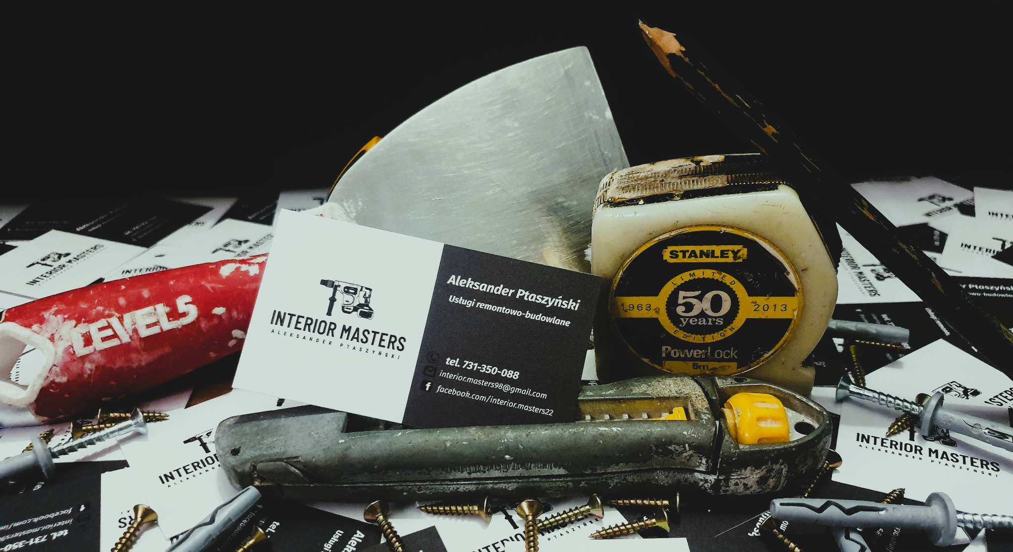 Narzędzia remontowe: szpachelka, miarka Stanley 50 years edition, ołówek, nożyk, kołki rozporowe i wkręty, wizytówki firmy budowlanej Interior Masters Aleksandra Ptaszyńskiego na czarnym tle.