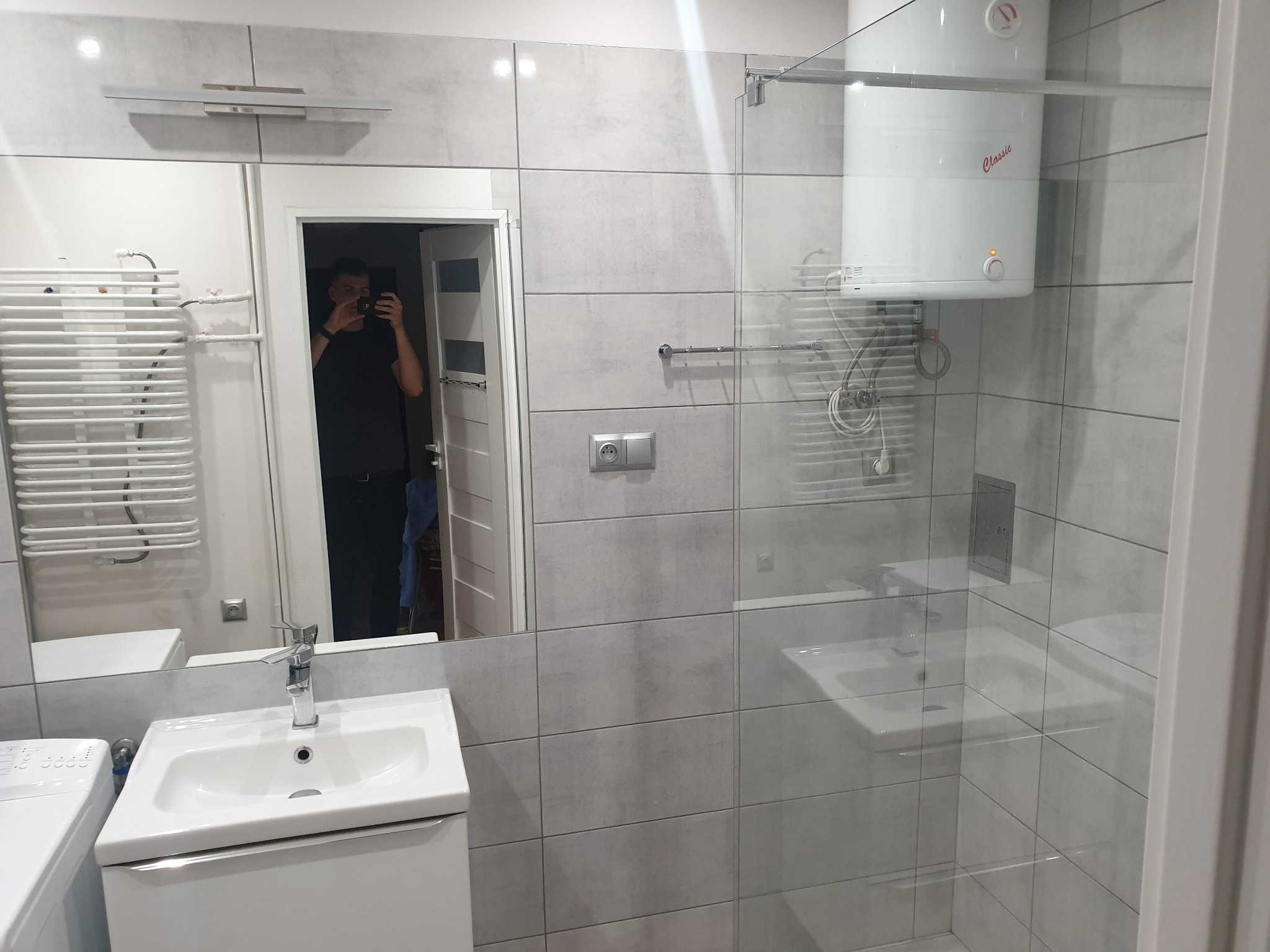 Wyremontowana łazienka z szarymi płytkami imitującymi beton, kabiną prysznicową ze szklaną ścianką, białym bojlerem oraz odbiciem fotografa w lustrze nad umywalką.