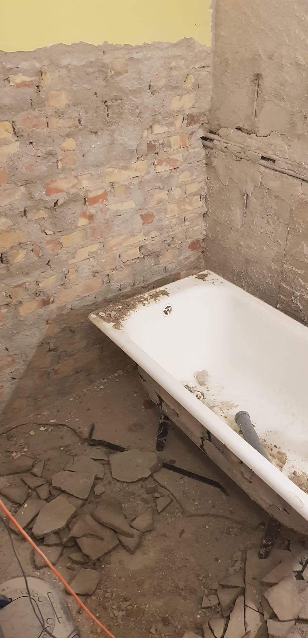 Biała wanna na metalowych nóżkach w trakcie demontażu w brudnej łazience z odsłoniętą cegłą, gruzem na podłodze i żółtą ścianą w tle.