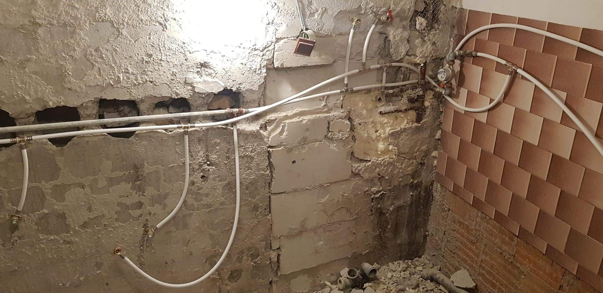 Fragment łazienki w trakcie remontu: odsłonięte instalacje wodne z białych rur na tle surowych ścian z widocznymi cegłami i resztkami starej glazury.