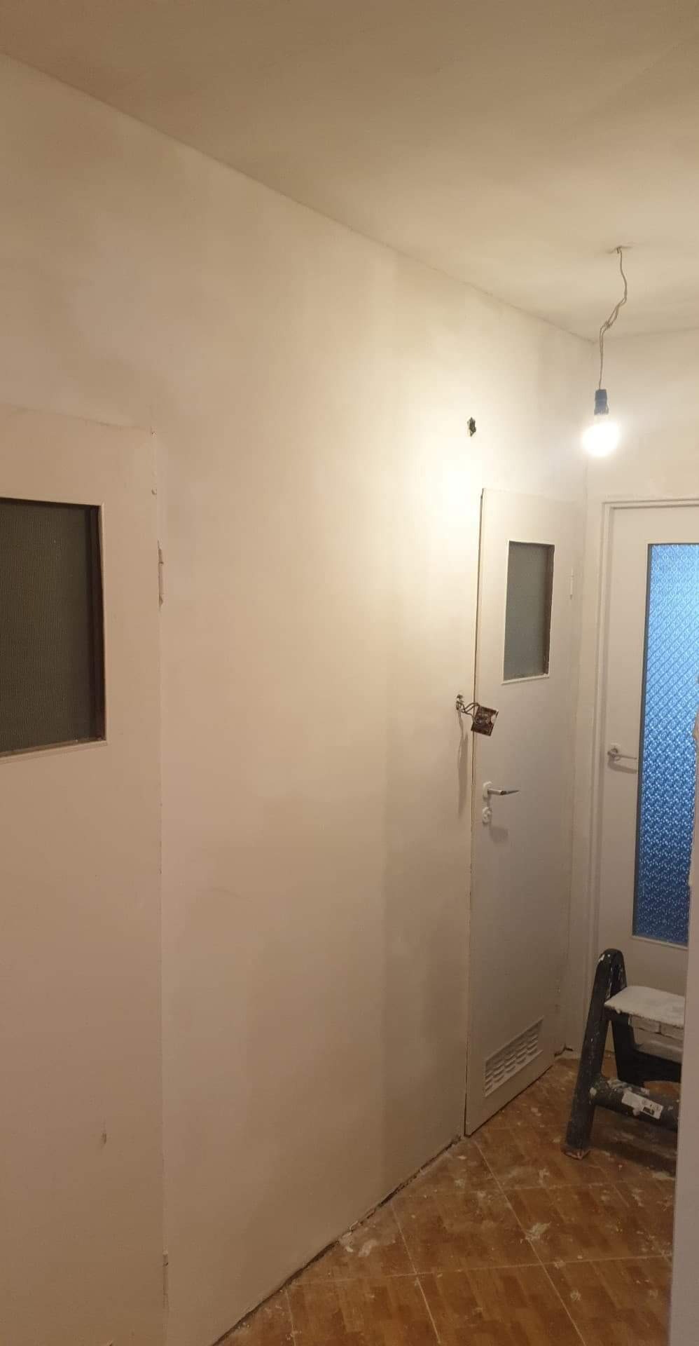 Korytarz w trakcie prac wykończeniowych: świeżo położone białe gładzie na ścianach, widoczne ślady po remoncie na podłodze, drabina oparta o ścianę obok drzwi z szybą, surowa żarówka pod sufitem.