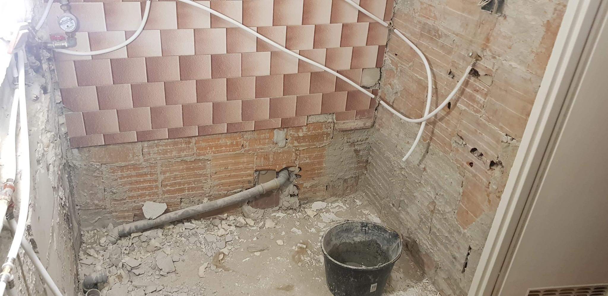 Łazienka w trakcie remontu: częściowo położone brązowo-beżowe płytki, odsłonięta cegła, rury, wiadro z zaprawą i gruz na podłodze.