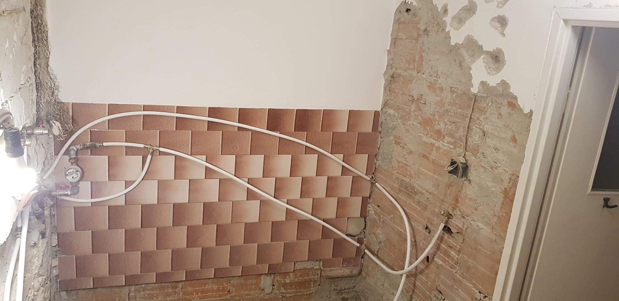 Fragment łazienki w trakcie remontu z częściowo ułożonymi, brązowo-białymi kaflami na ścianie, widoczne wystające rury instalacji wodnej i fragment gołej cegły z instalacją elektryczną.