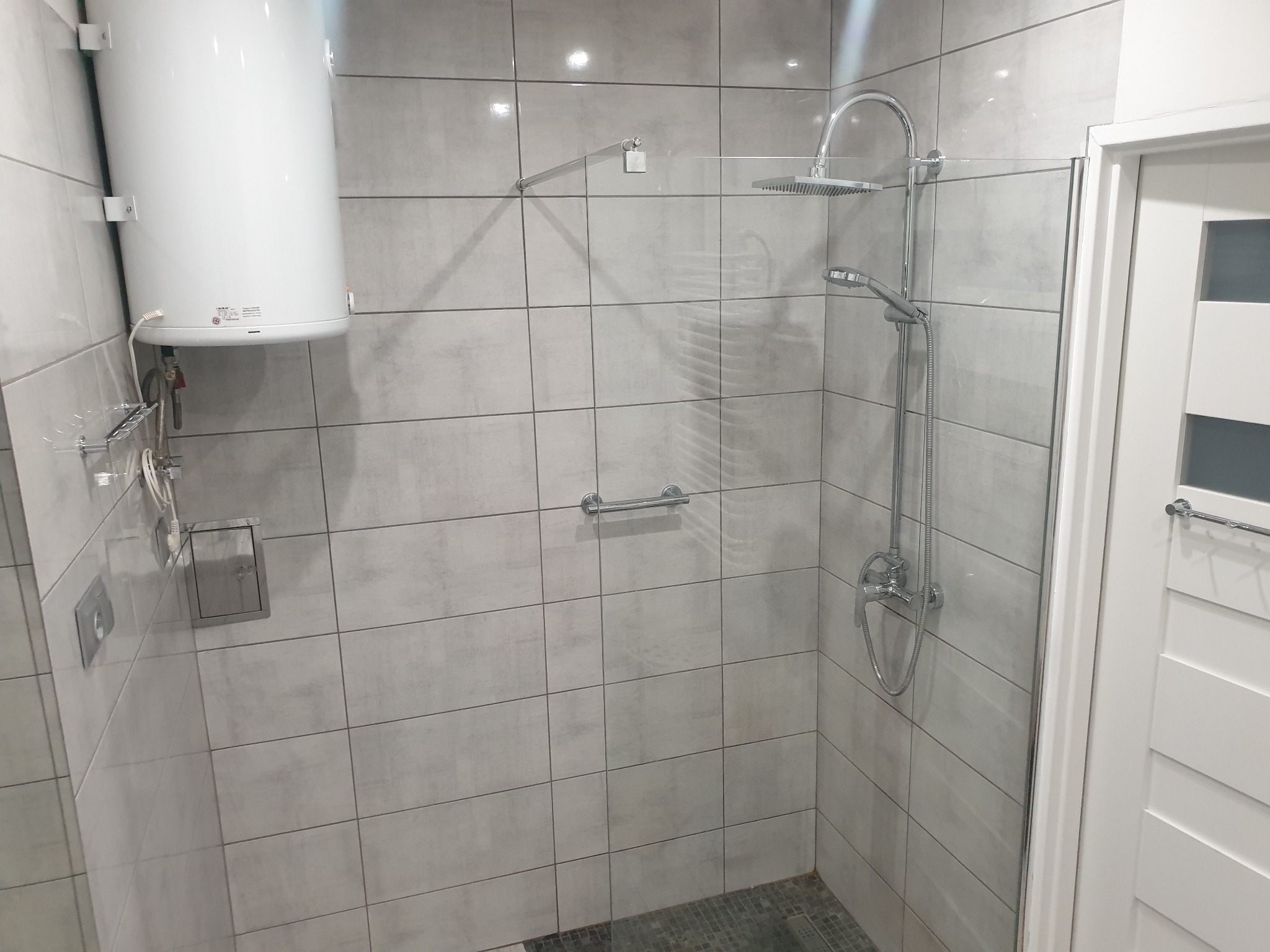 Nowoczesna kabina prysznicowa z deszczownicą i baterią termostatyczną, wykończona szarymi płytkami imitującymi beton, z widocznym bojlerem i uchwytem dla niepełnosprawnych.