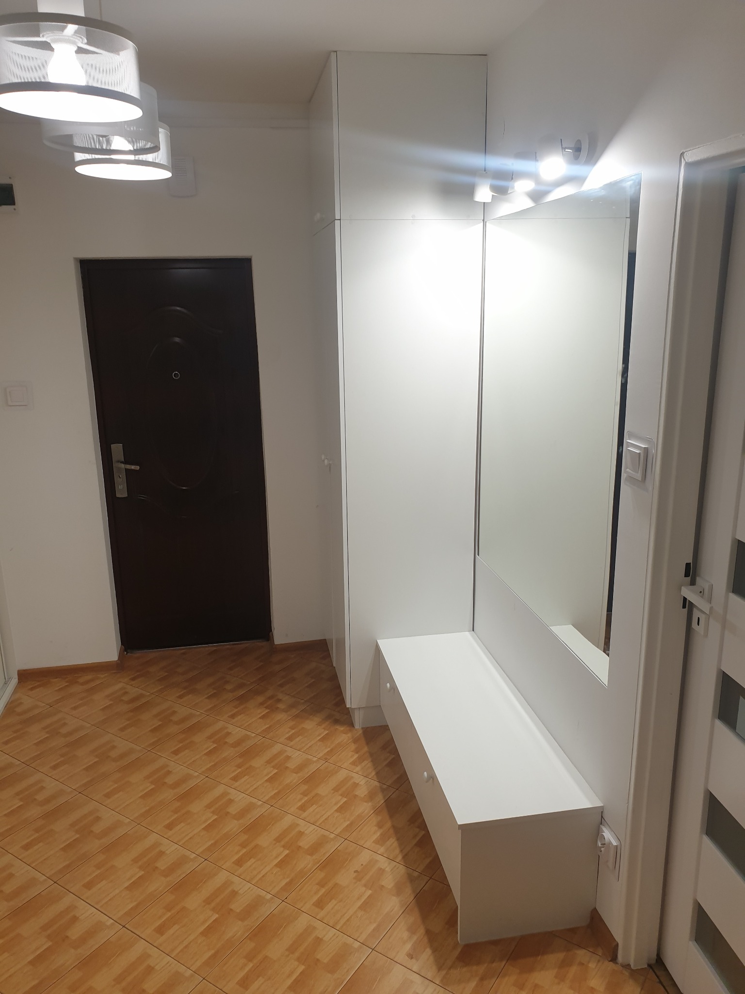 Nowoczesny przedpokój z zabudową w kolorze białym, obejmującą szafę, lustro i siedzisko, oświetlony trzema lampami sufitowymi z ażurowymi kloszami i dodatkowym oświetleniem punktowym nad lustrem...