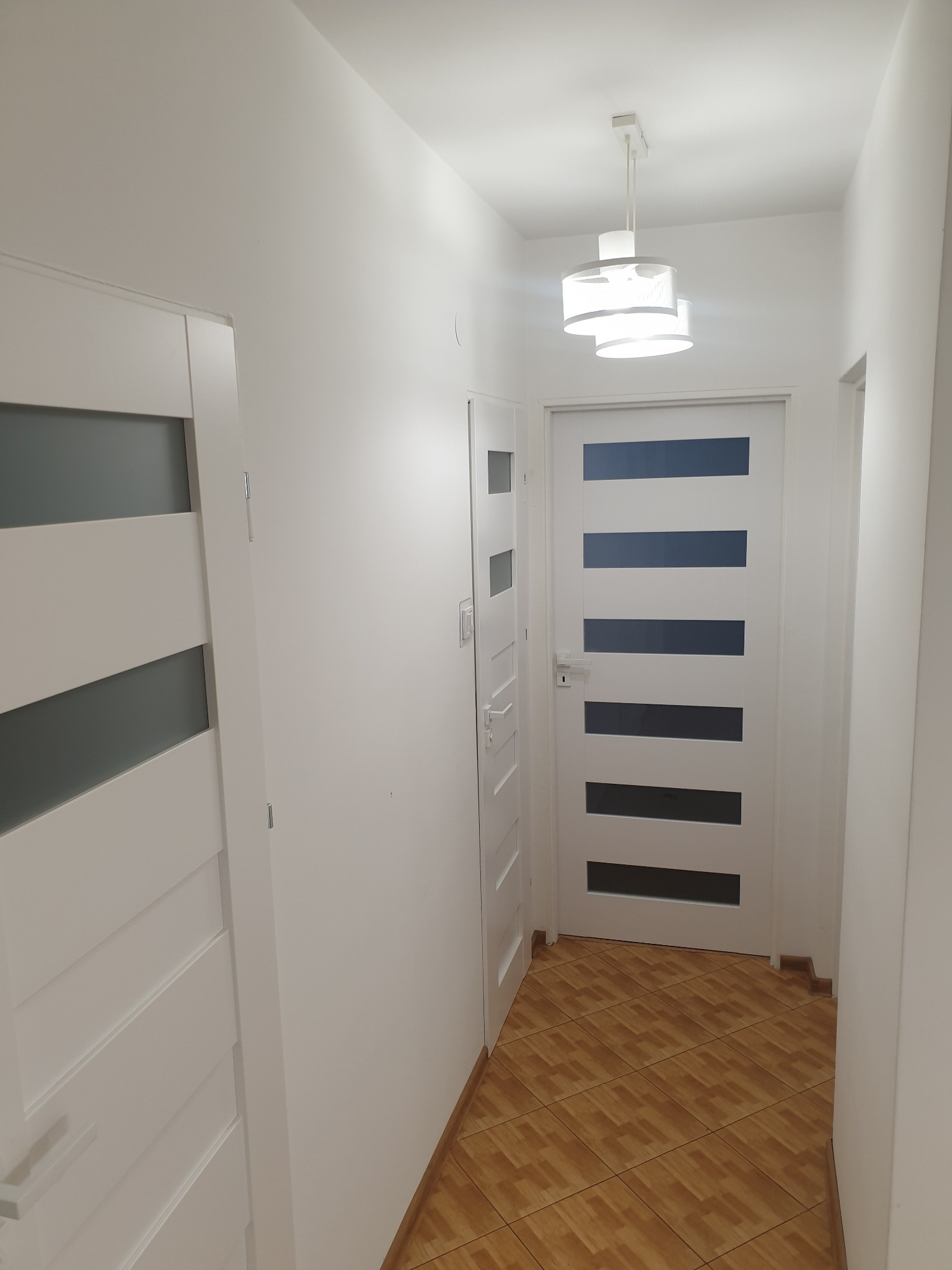 Wnętrze korytarza z dwiema białymi, nowoczesnymi drzwiami z przeszkleniami, oświetlonego lampą sufitową z białym abażurem i wykończonego brązowymi płytkami podłogowymi.