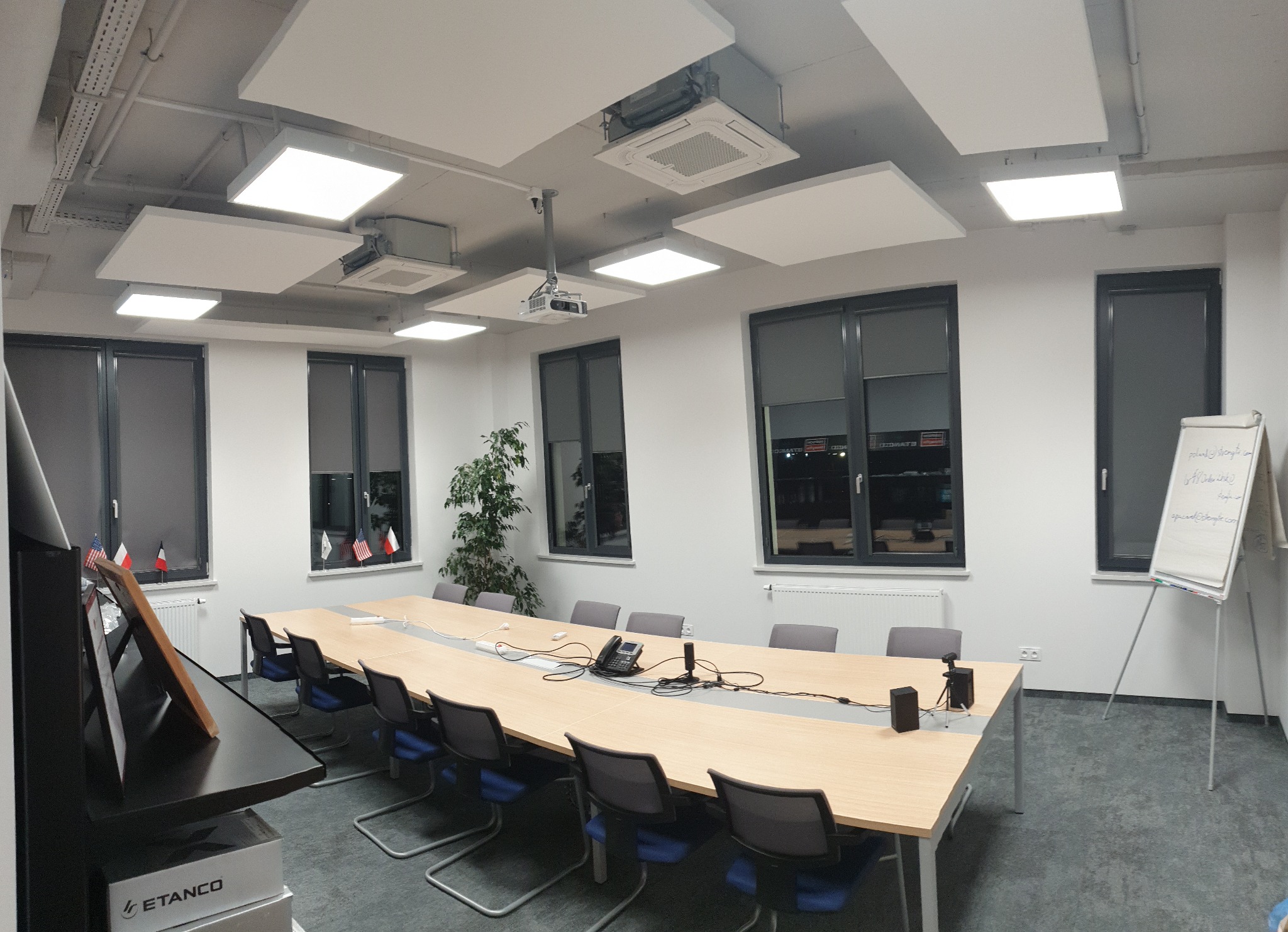 Sala konferencyjna z długim stołem, krzesłami, projektorem sufitowym, oświetleniem panelowym i tablicą suchościeralną, widok na okna z flagami
