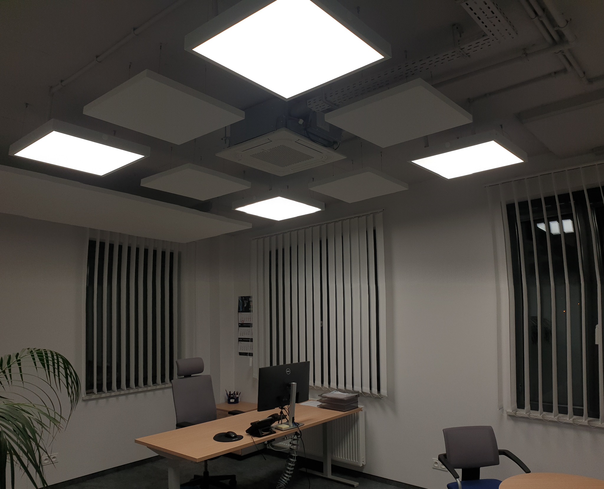 Biuro z podwieszanym sufitem z kwadratowymi panelami LED, widoczne biurka, krzesła, rolety pionowe i klimatyzacja sufitowa.